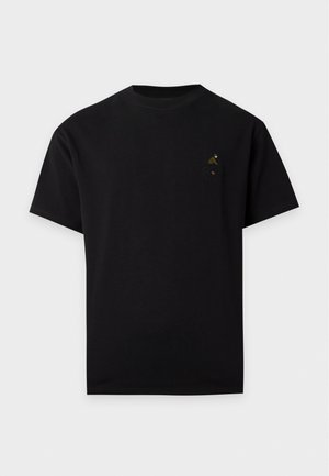 Un homme pose avec un T-shirt noir orné d’un petit emblème sur la poitrine, un jean gris ample et des chaussures marron, sur un fond clair uni.