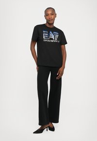 Sieviete, kura valkā melnu Emporio Armani EA7 sekvīnu logo t-kreklu, melnu platu bikses un melnas papēžu kurpes, stāvot pret baltu fona.