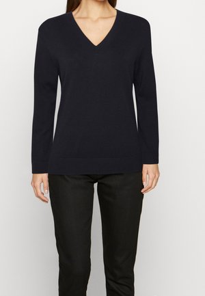 Žena nosící jednobarevný černý svetr s dlouhým rukávem a výstřihem do V a černé kalhoty stojící na neutrálním pozadí.