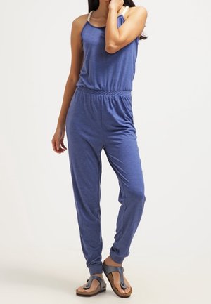Blauwe mouwloze jumpsuit met elastische tailleband en omgeslagen enkels, gemaakt van zachte stof. Draagt bruine sandalen met een teenlus.