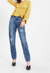 Haut jaune à manches longues côtelé avec un col rond et un détail plissé, associé à un jean en denim bleu déchiré et des ballerines noires à bouts pointus.