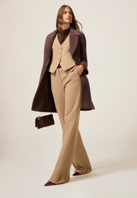 Bruine wollen jas over een beige vest en beige op maat gemaakte broek. Bevat een kantdetail, gouden knopen en rode puntige hakken met een bruine handtas.