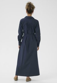 Robe chemise à manches longues bleu marine avec une taille ceinturée, détail de dos froncé et une silhouette droite. Fabriquée en tissu tissé, offrant une finition lisse.