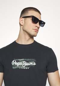 Camiseta negra con el logo blanco "Pepe Jeans London", de manga corta y textura suave. Gafas de sol negras con montura angular y audaz.