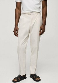 Světle béžové kalhoty s texturovaným, žebrovaným vzorem. Přiléhavý střih a sklady v přední části. Nosené s černými sandály. Minimalistický design.