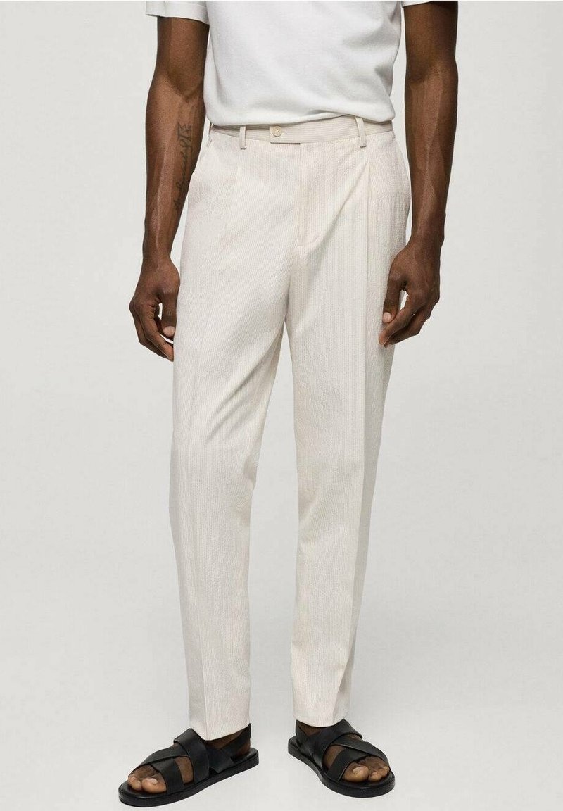 Světle béžové kalhoty s texturovaným, žebrovaným vzorem. Přiléhavý střih a sklady v přední části. Nosené s černými sandály. Minimalistický design.