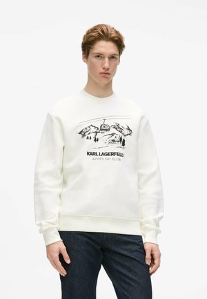 Junger Mann trägt weißen Pullover mit schwarzem Berg- und Skilift-Design und dem Text "Karl Lagerfeld Après Ski Club", kombiniert mit dunkler Hose.