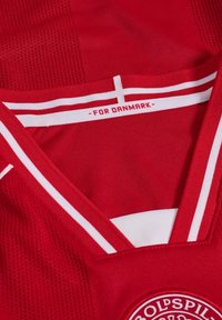 Colletto di una maglia sportiva rossa con strisce bianche, dettaglio di una croce bianca e testo "FOR DANMARK" sulla parte interna del collo.