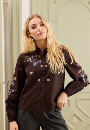 Jeune femme aux longs cheveux blonds porte une chemise marron foncé à manches longues avec des broderies florales bleues et beiges et un pantalon gris, debout à l'intérieur.