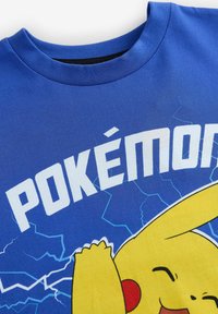 Zila kokvilnas T-kreklā ar Pikachu grafiku, baltiem uzrakstiem "POKÉMON" un grafiskām zibens akcentiem. Klasisks noapaļots kakls.