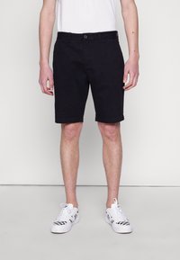 Sorte bomuldsshorts med lige snit, med knaplukning og sidelommer, parret med hvide sneakers med ternet mønster.