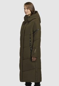 Lungo cappotto imbottito verde oliva con cappuccio, caratterizzato da un design trapuntato, tasche con zip e dettagli a bottone a pressione lungo i lati.