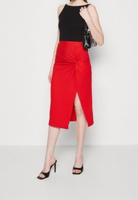 Haut sans manches noir associé à une jupe rouge mi-longue avec un détail twist et une fente, complété par des sandales à talons noires.