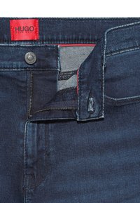 Tmavě modré džíny z denimu s částečně rozepnutým zipem, kovový knoflík s rytinou "HUGO BOSS" a červená etiketa přišitá uvnitř pásku.