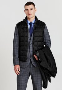 Schwarze gesteppte Weste mit Stehkragen über einem grauen karierten Anzug. Das Model hält eine schwarze Jacke, die ein schlankes Design und eine maßgeschneiderte Passform zeigt.