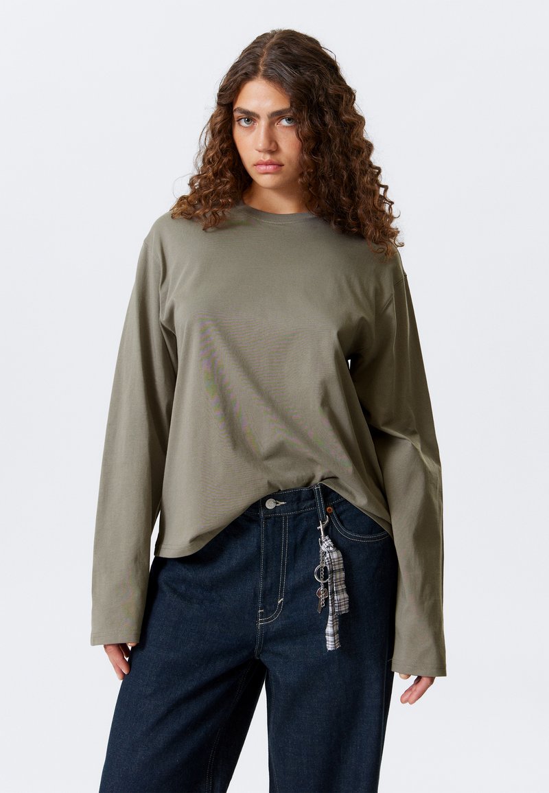 Olijfgroene langemouwen top van zacht materiaal, gecombineerd met hoge, donkerblauwe jeans met een decoratieve sleutelhanger en ruitpatroon.