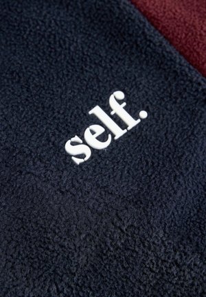 Tumesinine fliiskangas tekstuuriga viimistlusega; sellel on valget värvi kiri "self.". Samuti on nähtav burdgundi punane sektsioon.