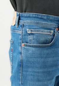 Blue denim jeans with a straight cut, featuring a leather patch on the waistband, copper rivets, and classic five-pocket styling.