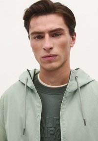 Lys grøn hættetrøje med en glat tekstur, lynlås lukning og justerbare snore, båret over en mørkegrøn sweater.