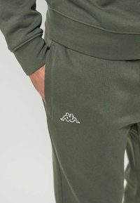 Persona che indossa pantaloni da tuta verde oliva con un logo bianco sulla coscia sinistra, mano appoggiata casualmente nella tasca.