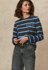 Langærmet gråstribet skjorte med lyseblå vandrette striber. Har "BOSTON" broderet i blåt. Kombineret med højtsiddende mørke jeans.