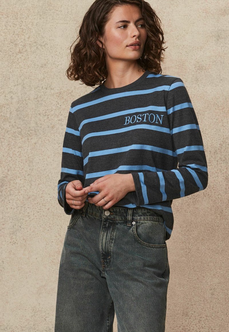 Camisa de manga larga a rayas grises con rayas horizontales en azul claro. Presenta "BOSTON" bordado en azul. Combinada con jeans oscuros de cintura alta.