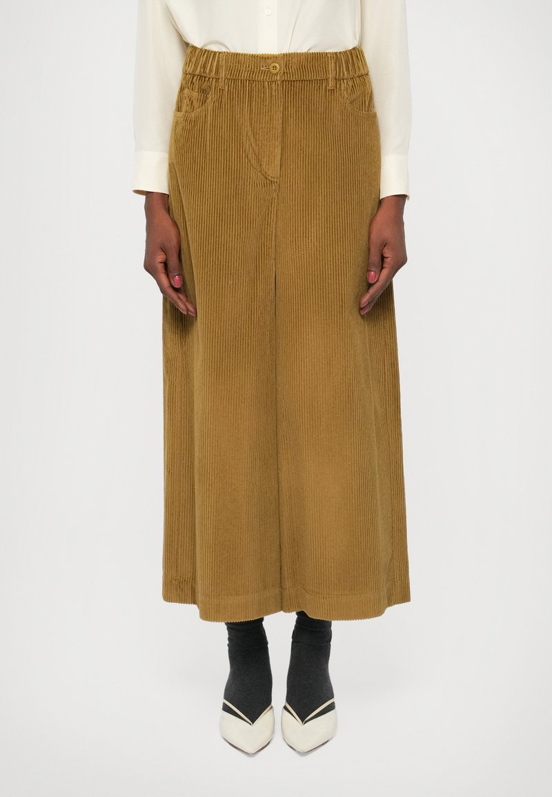 Pantalones de pana marrón de pierna ancha con cintura elástica abotonada, bolsillos laterales y un patrón acanalado texturizado. Combinados con calcetines negros y zapatos de punta.