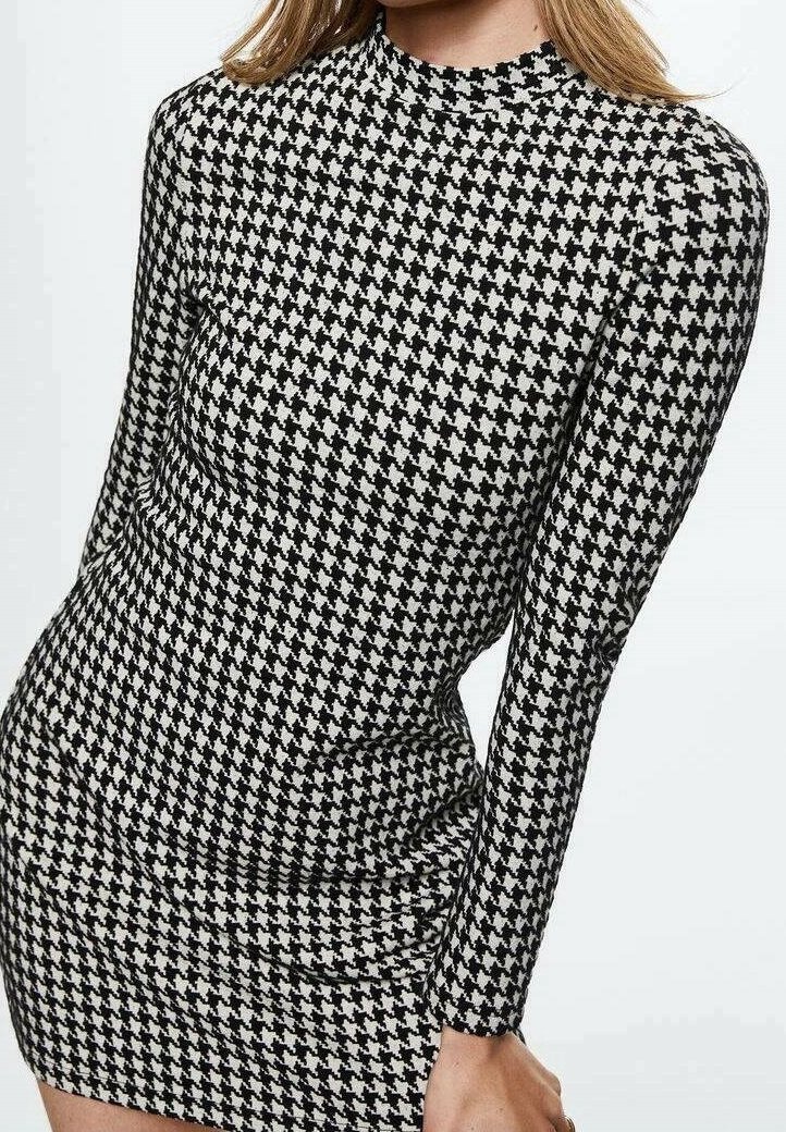 Femme portant une robe cintrée à manches longues, avec un motif pied-de-poule noir et blanc, debout devant un fond clair uni.