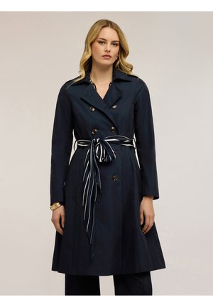 Donna che indossa un trench doppiopetto blu navy con cintura a righe annodata, in piedi davanti a uno sfondo chiaro uniforme.