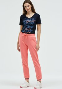 Marineblaues T-Shirt mit grafischem Schriftzug, kombiniert mit korallfarbenen Jogginghosen mit elastischem Bund und Bündchen. Weiße Sneakers mit rosa Akzenten.