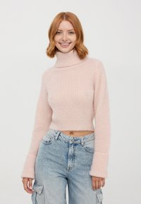 Maglione rosa chiaro a coste con collo alto e maniche larghe, abbinato a jeans cargo blu scoloriti. Texture morbida e design accorciato.