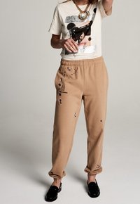 Beige Jogginghosen mit elastischem Bund sind mit Farbspritzern verziert. Kombiniert mit einem T-Shirt mit Grafikdruck. Schwarze Loafers runden den Look ab.