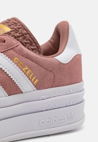 Sapatilhas em camurça rosa com detalhes em couro branco, logótipo dourado "Gazelle", parte superior texturizada e uma sola branca grossa com branding em relevo.