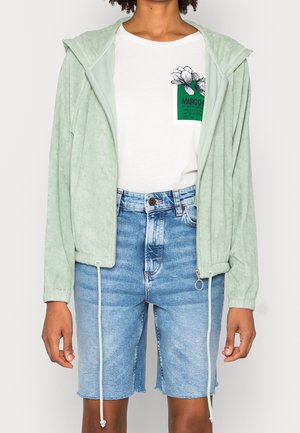 Person trägt eine hellgrüne Kapuzenjacke mit Reißverschluss, ein weißes T-Shirt mit Blumenmotiv und Text sowie hoch taillierte hellblaue Jeansshorts.