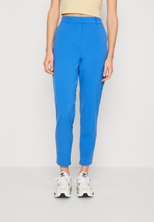 Pantalons bleu sur mesure avec une texture lisse, coupe slim, longueur raccourcie et poches latérales, associés à des baskets blanches et beiges.