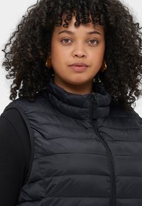 Juodas pūkuotas liemenės su aukštu apykakle, su horizontaliai kanaluotomis dalimis ir priekiniais užtrauktukais. Lygi tekstūra ir prigludęs dizainas.