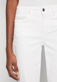Jeans in denim bianco con un design aderente, caratterizzati da una tasca laterale decorata con una striscia di strass argentati e chiusura con bottone metallico standard.