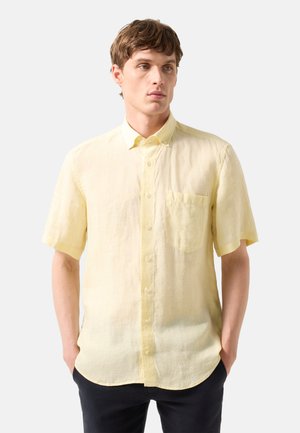 Jonge man met kort bruin haar, dragen een lichtgele korte mouwen overhemd met borstzak en zwarte broek.