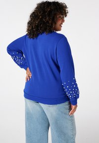 Blauwe sweater met parelversieringen op de mouwen. Heeft een ontspannen pasvorm en een geribbelde zoom, gecombineerd met lichtblauwe wijdlopende jeans.