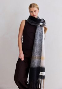 Écharpe douce et surdimensionnée en gris, noir et crème, avec de subtiles touches de jaune et des détails à franges. Fabriquée en tissu texturé et chaud.