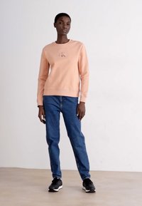 Sudadera rosa con cuello redondo, jeans de mezclilla azules y zapatillas negras. La sudadera presenta un pequeño logotipo en el pecho.