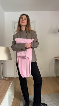 Γυναίκα με γκρι σακάκι και μαύρο παντελόνι κρατά διπλωμένη ροζ φόρμα, στέκεται σε ένα φωτεινό δωμάτιο με ξύλινο πάτωμα και λευκούς τοίχους.