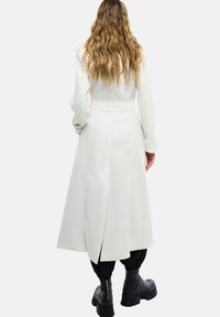 Manteau long blanc en tissu lisse et texturé avec une taille ceinturée, doté d'une fente à l'arrière et associé à des bottes noires zippées.