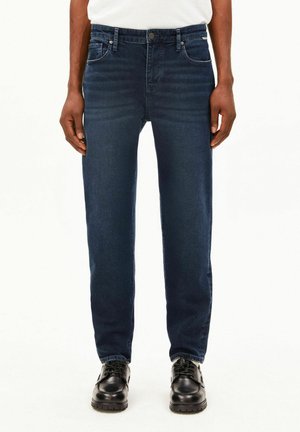 Person trägt dunkelblaue Jeans, ein weißes Hemd und schwarze Schuhe und steht aufrecht vor einem schlichten weißen Hintergrund.