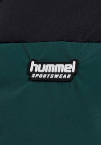 Hummel Sportswear-logotypen på ett färgblockerat tyg som har mörkgröna och svarta sektioner med en texturerad yta och formpressade gummidetaljer.