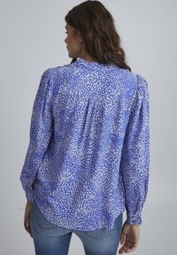 Blue patterned blouse with white speckles, long sleeves, gathered shoulders, and a curved hem. Fabric appears lightweight and smooth.