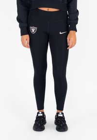 Zwarte leggings met een Oakland Raiders-logo en een witte Nike swoosh. Rekbaar materiaal met een aansluitend ontwerp, gecombineerd met zwarte sneakers.