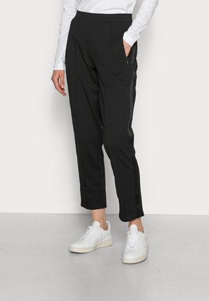 Person wearing black ankle-length pants with side stripes, white sneakers, and a white long-sleeve shirt, standing against a white background.