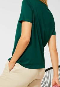 Mörkgrön kortärmad t-shirt med avslappnad passform, mjukt tyg, rundad nederkant och rund hals, matchad med ljusbeiga byxor.