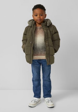 Olivgrüne Pufferjacke mit einer grauen Fleece-gefütterten Kapuze, getragen über einem gradienten Strickpullover, mit blauen Jeans und weißen Sneakers.